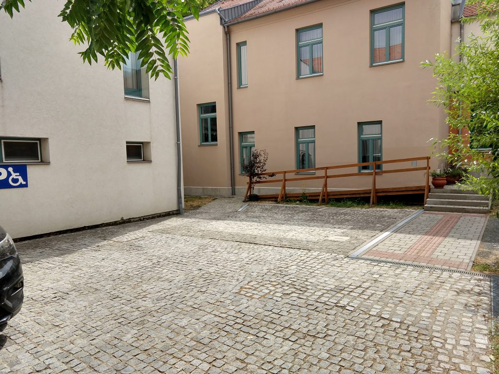 Két épület közti udvar. Balra egy magas, fehér épület látható. Az épület előtt fekete autó parkol, mellette a falon egy kék, mozgássérült parkolót jelző tábla. A szemben levő épületig széles, kövezett járda. Ez az épület bézs színű, rajta 2 sor, türkíz keretű ablak. Az épülettől balra egy rámpa kezdődik, mely az épület végéig vezet. A rámpától jobbra fakorlát. Az épülettől jobbra, kicsit beljebb található a bejárat, melyhez 4 lépcső is vezet, a rámpa helyett használható. A bejáratot eltakarja egy fa, nem látjuk jól.