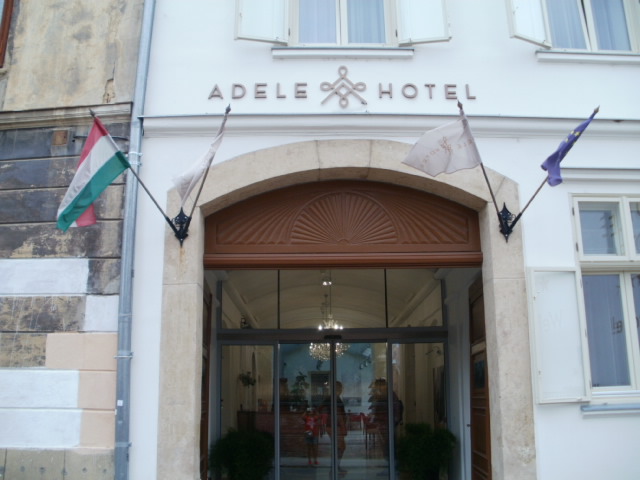 Kapubejáró utcáról. A felirat a kapu felett, ADELE HOTEL. Modern és a történelmi keveredése. A bejárat mögött pár méterrel fotocellás üvegajtó látható ami a recepcióhoz vezet. Két oldalt cserepes növények. A kapu két oldalán két-két zászló van, magyar nemzeti,Európai Uniós. és szálloda sajátja.