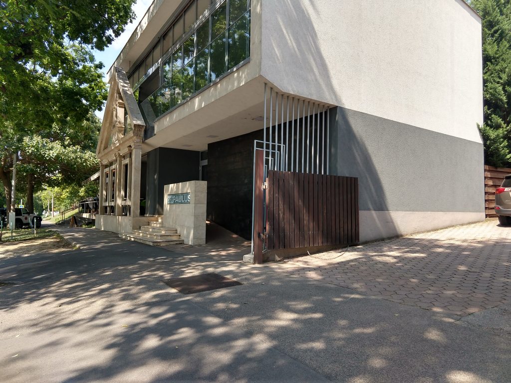 Modern épület amely ötvözi a minimalista és klasszikus elemeket. Az épület elején 5 lépcső, mellette egy félfal PAULUS felirattal, a félfal mögött rámpa. A ház jobboldalán gépkocsibejáró kapuval, elötte árnyat adó fák sora.