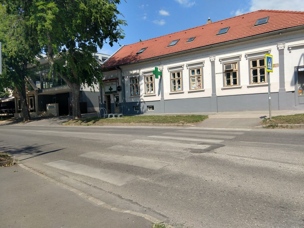 Utcarészlet, zebrával. A gyalogosátkelő mindkét végén rámpával. Szemben egy polgári jellegű ház, melynek egyik végében gyógyszertár található. Az háztól balra egy modern épület, elötte két árnyat adó fával.