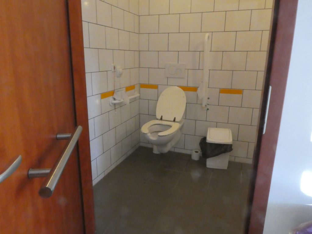Az akadálymentes WC szabályosan kétoldalán kapaszkodókkal. A fal felőli oldalon a falra rögzítve fix, a másik oldalon felhajtható. A WC csésze azonban szabálytalanul nyitott elől, ami balesetveszélyes. A WC mellett nagy kuka látszik.