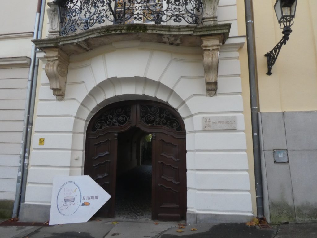 A Szent István tér felőli bejárat. Régi gyönyörű épületet látunk, boltíves fa nagykapuval. Fölötte kis erkély kovácsoltvas korláttal.