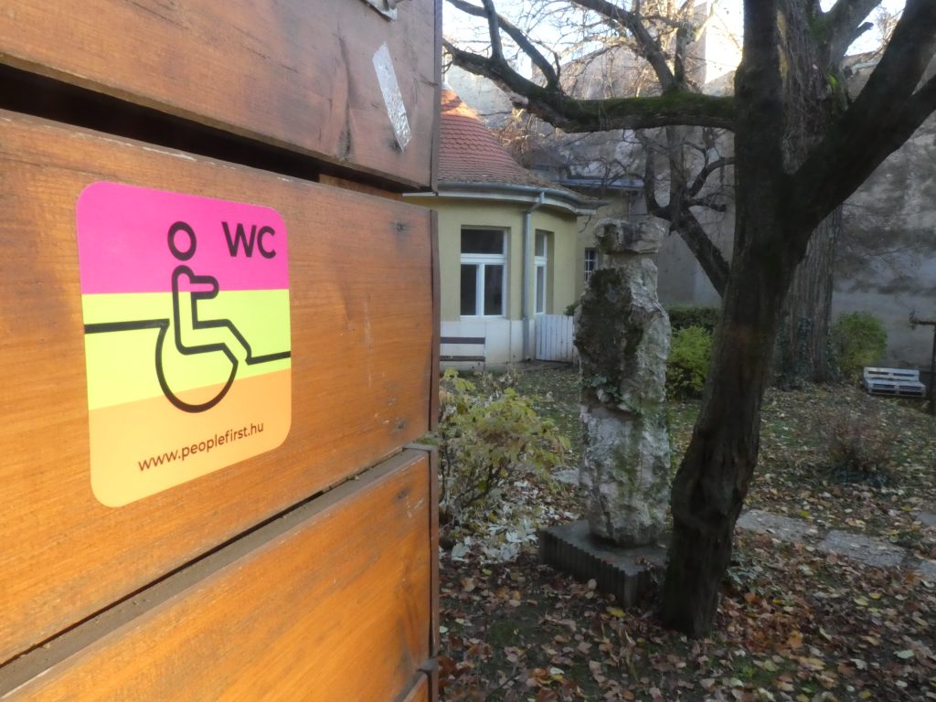 Az akadálymentes bejárat a kert felől, a képen közelről látszik az egyesület matricája, háttérben a szép épület egy része, illetve a park bokrai, fái.