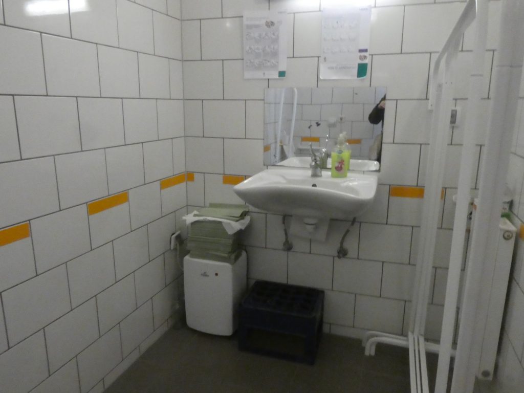 Az akadálymentes WC kézmosóját fotóztuk. Szabályosan íves, mellette balra a vízmelegítő kisbojler a földön. Jobbra sajnos a kép készültekor éppen fém állványokat pakoltak be, ezek kikerülhetőek, de balesetveszélyesek.