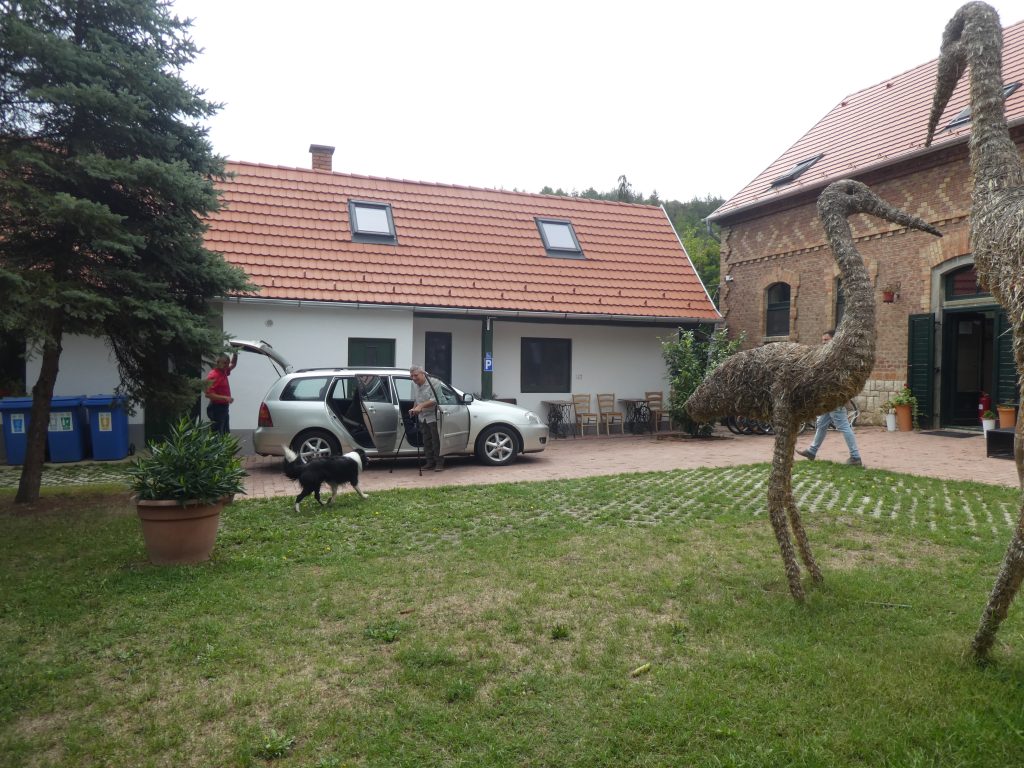 Az akadálymentes parkoló az udvar felől. Az udvarban dekorációs, nagyméretű szalma madarak, szemben egy kisház, jobbra a recepció pajtaépülete. A kisház előtt tégla burkolaton áll egy autó, ez az akadálymentes parkoló, a recepciótól mintegy két méterre lehet megállni.