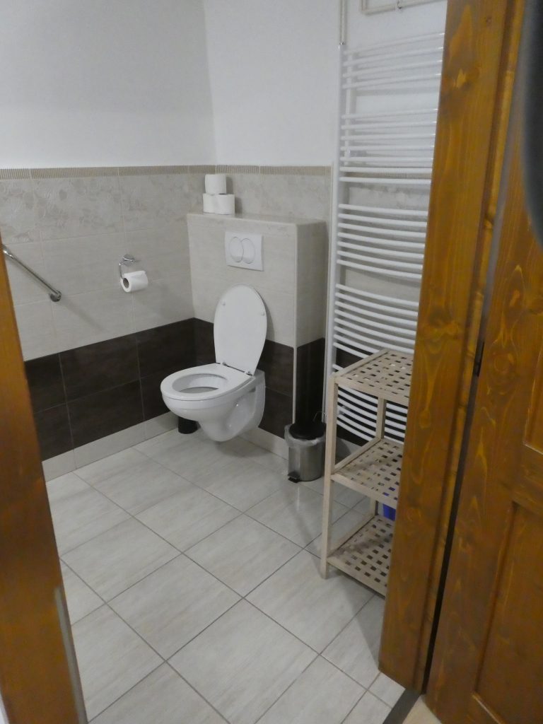 Az akadálymentes apartman WC-je. A fürdőbe belépve jobbra szemben a sarokban található. Szabályszerű kapaszkodók nincsenek, a WC-n ülve jobb oldalon a falon találunk egyetlen fém kapaszkodót. A burkolat világos csempe mindenütt.