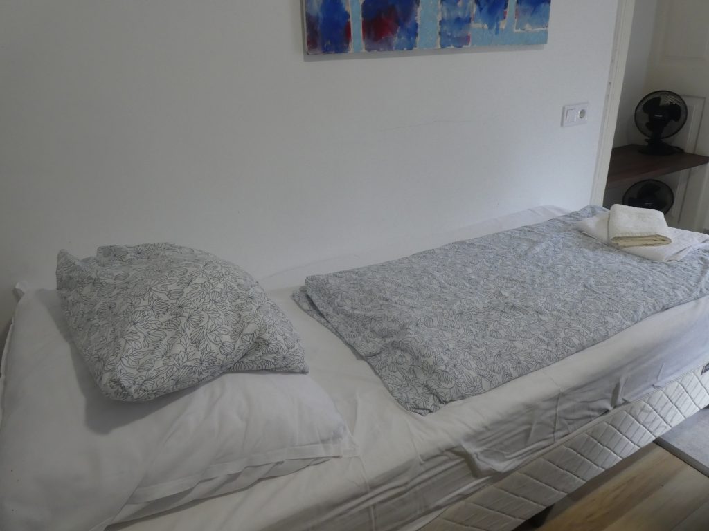 A hálóban az egyik ágyat látjuk. Viszonylag keskeny, egyszemélyes ágy, magas, kényelmes matracokkal. Az ágy fölött képpel.