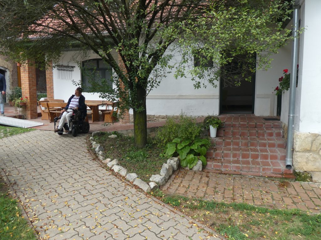 Az akadálymentes apartman kintről. Nagyon szép hangulatos bejárat 5 tégla lépcsővel szemből, vagy baloldalról épített, kis lejtős rámpával megközelíthetően. Jobb oldalán a házból kiemelkedő félig zárt teraszrész látható. Szemben a bejárati ajtó. Balra a rámpán lejőve egy kis beugróban fa asztallal és padokkal berendezett kellemes teraszrész az udvar szintjén.