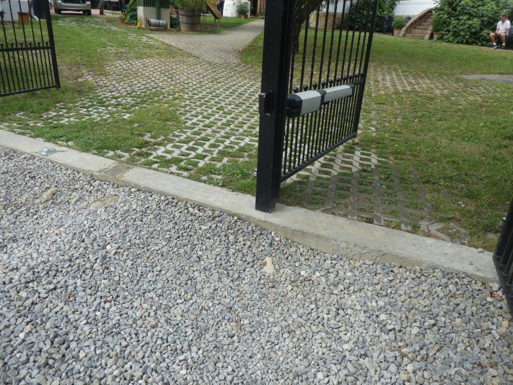 A durva murvás utcai bejárat közelebbről. Itt jól látható, hogy a murvás rész és a kapu között a kb. 10 cm-es küszöb is található a gyalogos bejáratnál. Utána térkővel lerakott udvarrész folytatódik, a térkövek lyukacsosak, közöttük kinő a fű.