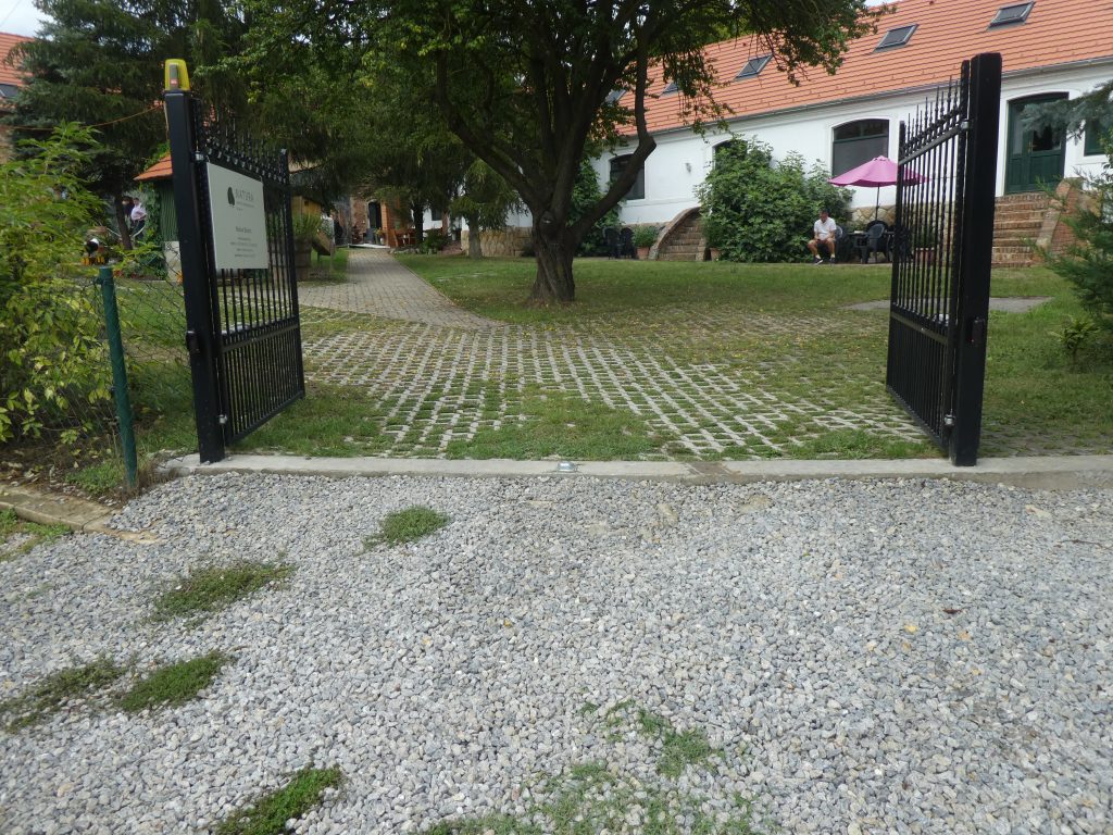 Az utcai bejárat a panzió parkjába. Durva murvás felületet látunk, a nagy kapu baloldali része nyitható az autóknak, jobb oldali része a gyalogos bejárat. Szemben a panzió L alakban látható, szemben a főépület, jobbra két apartman.