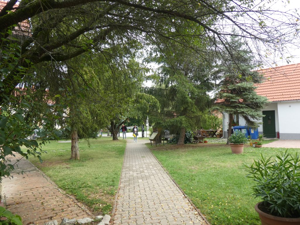 A panzió udvarát látjuk ismét, az utcáról belépve. Két irányba vezet térkőből kirakott egyenes, sima járda, balra az akadálymentes parkolóhoz és egyben a recepcióhoz, egyenesen a főépülethez, ami tulajdonképpen szintén a recepcióhoz vezet, nincs nagy távolság sehol. A park fákkal, bokrokkal, füves területtel nagyon kellemes.