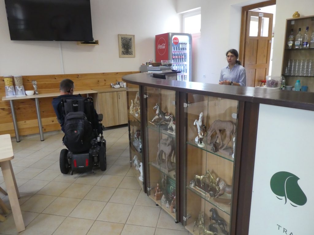 A recepció mögött a dolgozó hölgy, előtte Balázs kerekesszékben. A recepció pultja Balázs feje fölött található. A recepciós pult elülső oldala üvegből készült, dekorációs tárgyakat mutat.