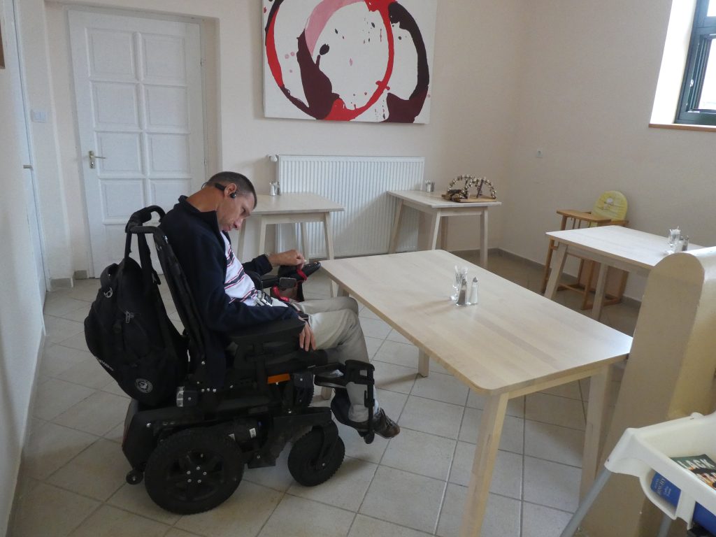 Balázs kipróbálja az egyik ebédlőasztalt, befér a lába az asztal alá kerekesszékkel is. Az ebédlő két részre van osztva, a bejárati rámpa felezi meg. Egyik felében négy négyszemélyes faasztal székekkel, másik felében szintén 3 asztal és magasabb bárasztalok találhatóak, ebben a felében van a recepciós pult is. Világos, tágas, kellemes terem. A fal mellett több helyen gyerek etetőszékek várnak használatra. A falakon nagyméretű absztrakt képek.