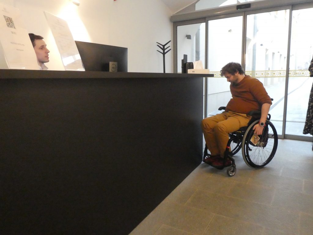 A recepció pultja mellett áll Gergő kerekesszékben. A pult a feje fölött kezdődik. Túl magas.