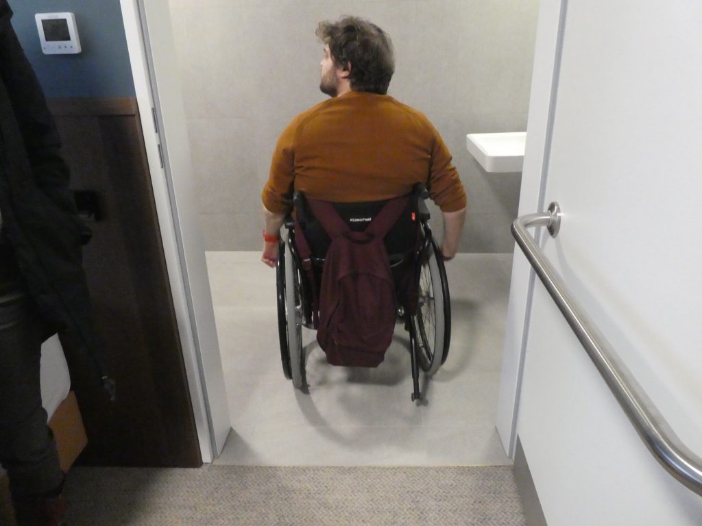 Az akadálymentes szoba fürdőszoba bejáratában áll Gergő kerekesszékkel. Küszöb nincsen.