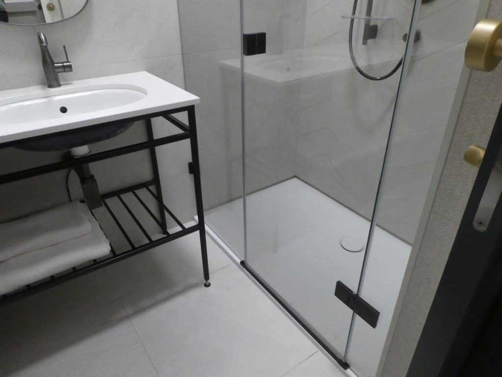 A hotel nem akadálymentes szobájában a tusolókabin. Az egész falat betölti, üvegajtóval nyílik, a tálca minimális (kb.1 cm) magasságú.