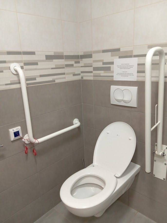 A WC közelről: a falon L alakú kapaszkodó, a másik oldalon felhajtott kapaszkodó. A WC szabályosan akadálymentes. Vészhívó az L alakú kapaszkodó alatt található.
