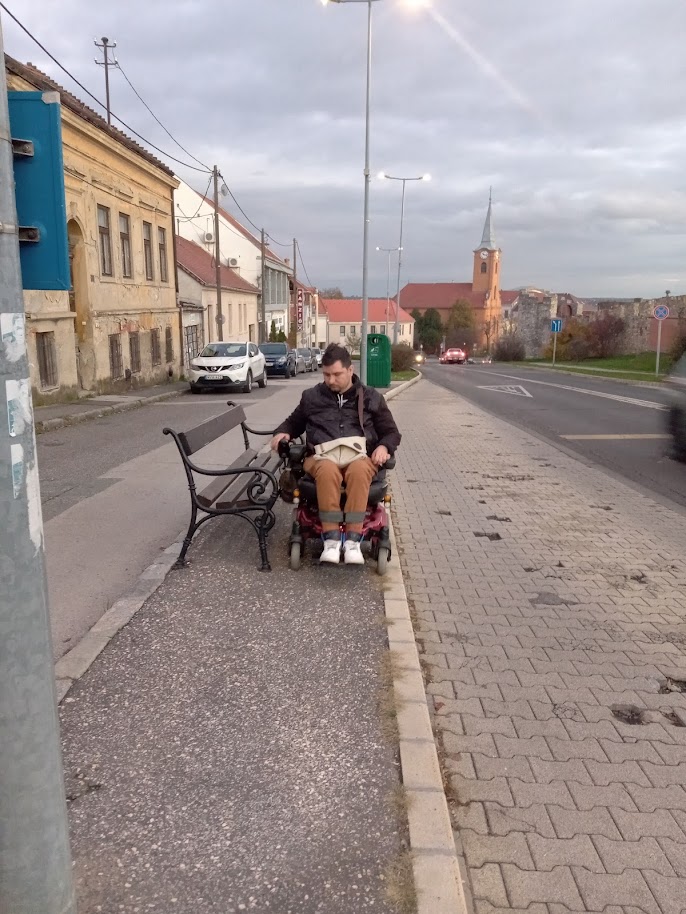 A buszmegállóban a járda közepén egy pad nehezíti a közlekedést. E mellett áll Roland kerekesszékkel, mutatva, hogy épp, hogy elfér a pad mellett.
