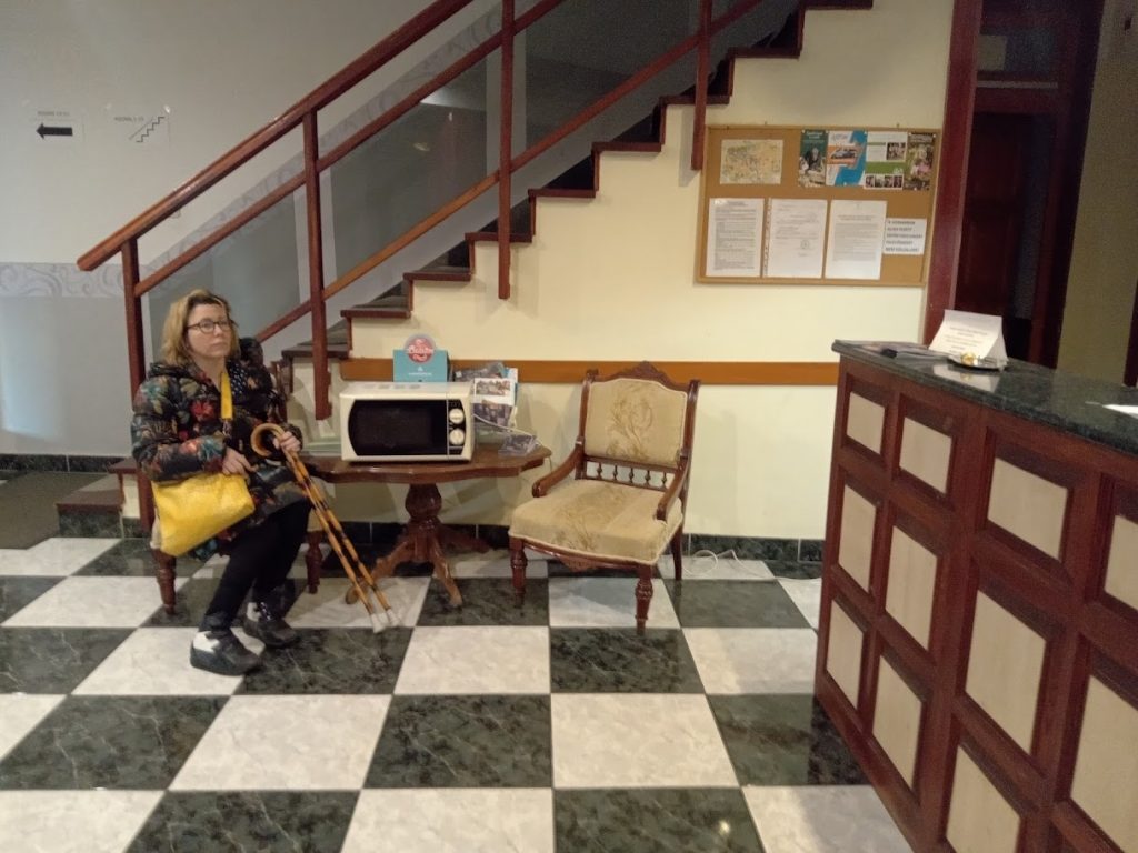 A recepció előtt ül Gyöngyi. Kontrasztos, kockás kő padlóburkolat, a recepció mellmagasságú, előtte egy kisasztal két régi stílusú székkel.