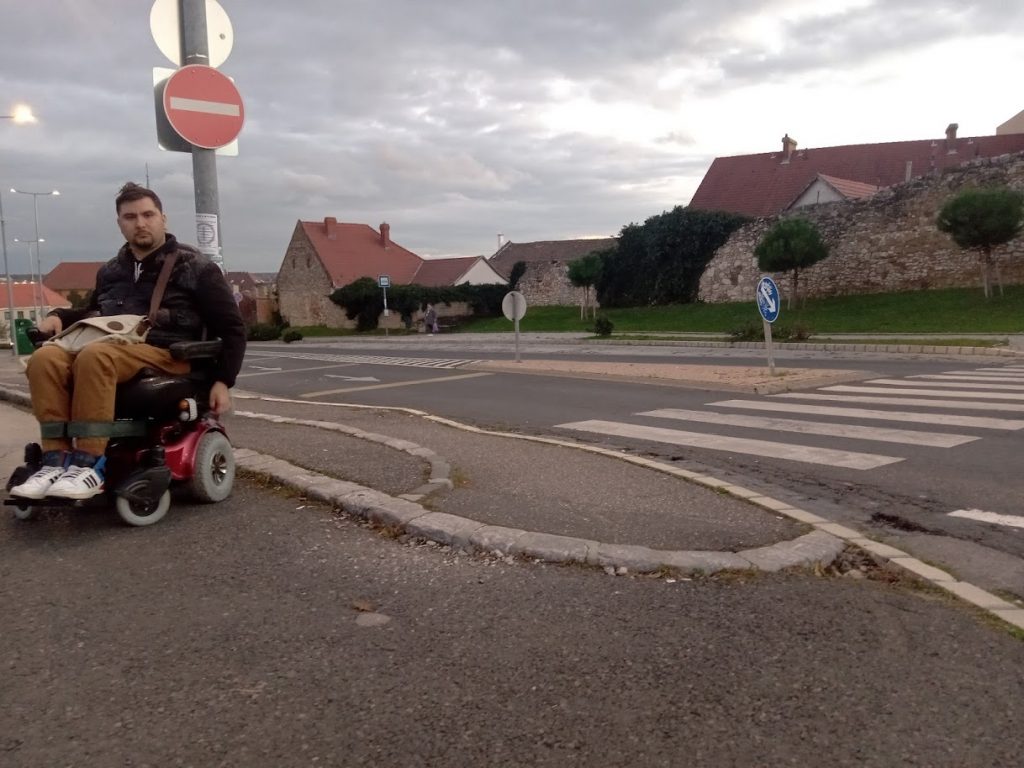 A járda végét látjuk a Várfal panzióhoz vezető zebránál. Lekerekített, akadálymentes. A képen Roland áll a járdán kerekesszékkel.