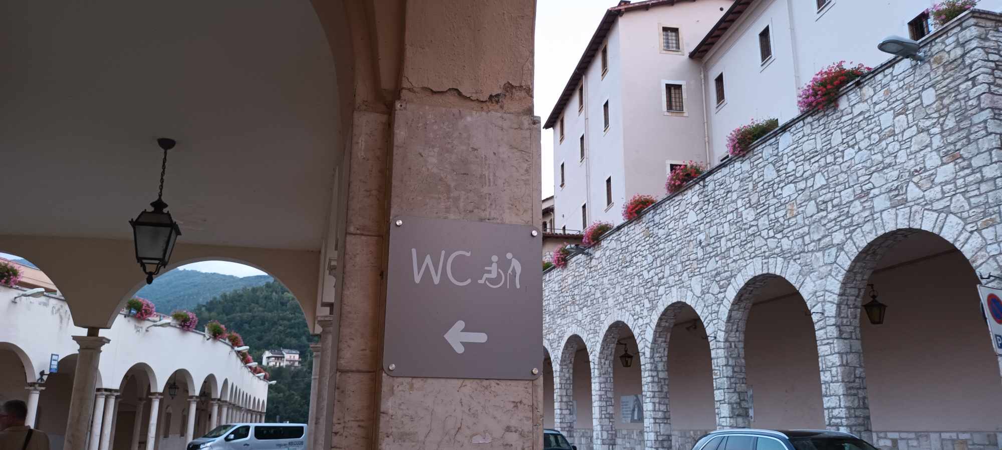 Jobboldalon régi árkádos épületet látunk, baloldalon szintén árkádok alatt a falon egy jelzés az akadálymentes WC-t mutatva.