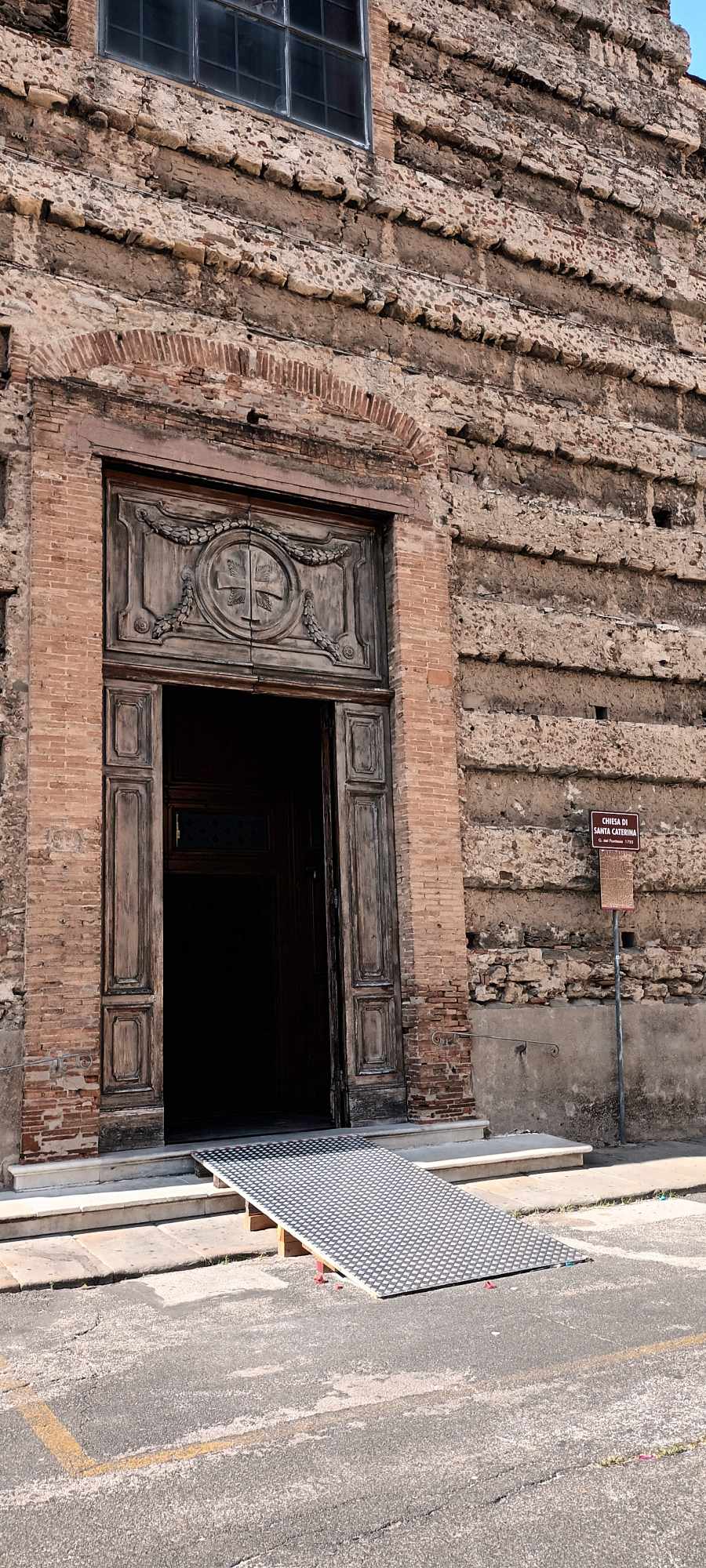 Nagyon régi templom bejáratát és falát látjuk. Az ódon fa kapu előtt lépcsők, rajta azonban kellemes lejtésű rámpa.