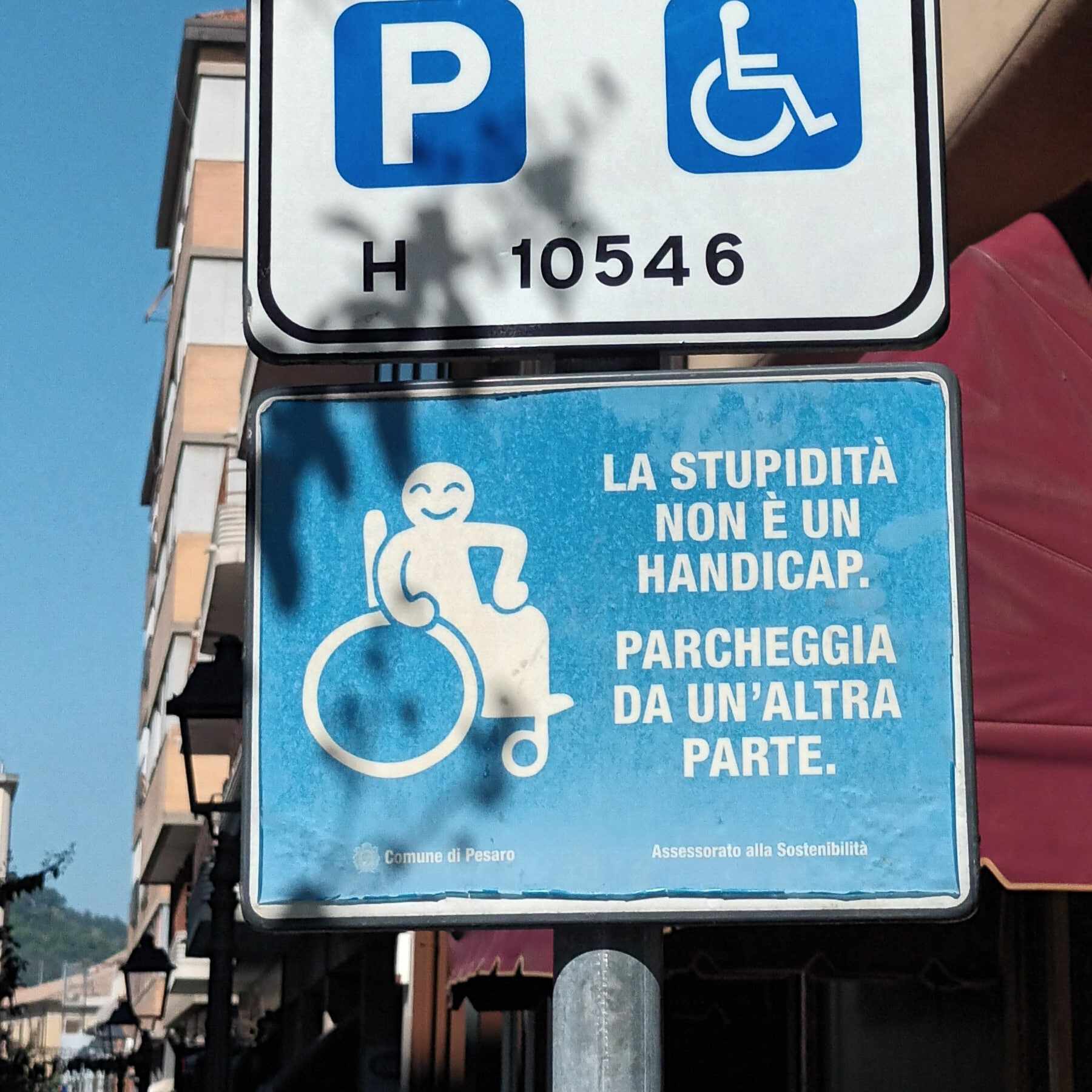 Az utcán egy parkolótáblát látunk. A hivatalos akadálymentes parkolót jelző tábla alatt egy vicces tábla egy nevetős kerekesszékes ikonnal és a szöveggel olaszul: "A hülyeség nem fogyatékosság. menj és parkolj máshova"