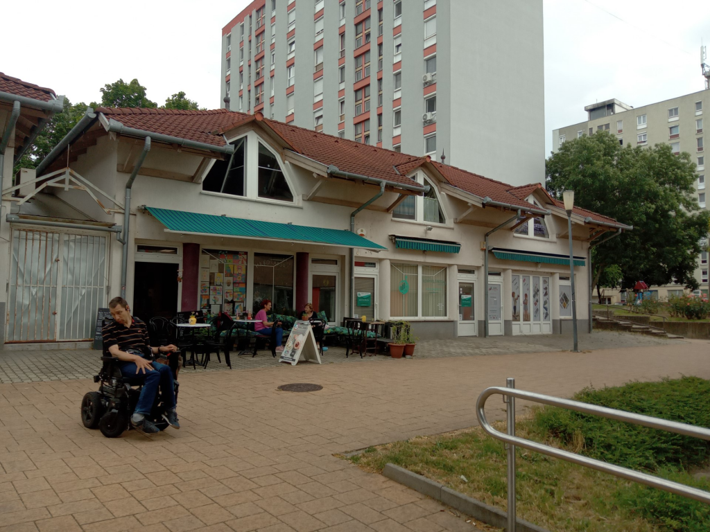 Kívülről látjuk a helyszínt. Egy üzlet a sok közül, viszonylag nagy tér előtte. A teraszon vendégek üldögélnek, az előtérben Balázst látjuk kerekesszékben, a rámpa tetején áll.