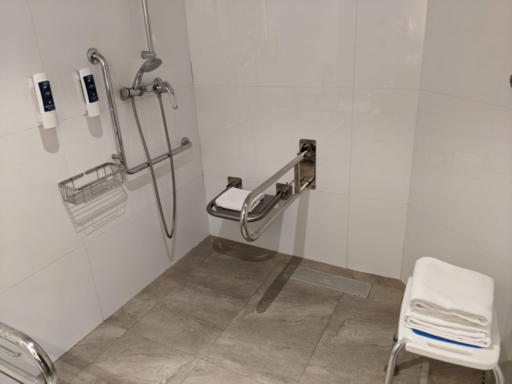 Akadálymentes fürdőszoba, lehajtható zuhanyülőkével és kapaszkodóval. A falon egy L alakú kapaszkodó és sampon valamint tusfürdő adagoló. Fehér csempe oldalfalakkal és természetes mintájú padlóval.
