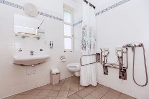 Egy fürdőszoba belső, mely teljesen akadálymentesen felszerelt. Szemben kézmosó, mellette a WC, kapaszkodóval, emellett zuhanyfüggönnyel elválasztva egy szintén kapaszkodókkal, ülőalkalmatosságokkal felszerelt tusoló. A padló sima csempés. 