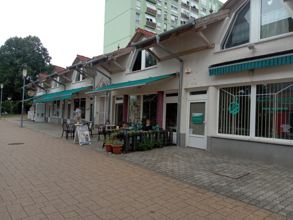Kívülről a hely. Sima beton burkolat található az épület előtt. Sok kisebb üzlet között helyezkedik el a pub, a panelházak előtt.