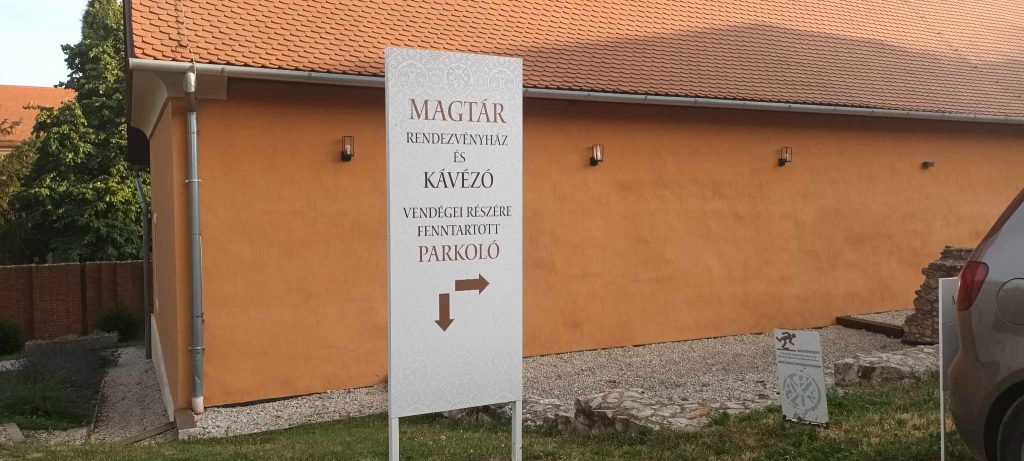 A parkolót jelző tábla. Felirata: Magtár rendezvényház és kávézó. Vendégeink részére fenntartott parkoló. És két nyíl jobbra (oldalsó parkolóhoz) és balra a Magtár előtti parkolóhoz.