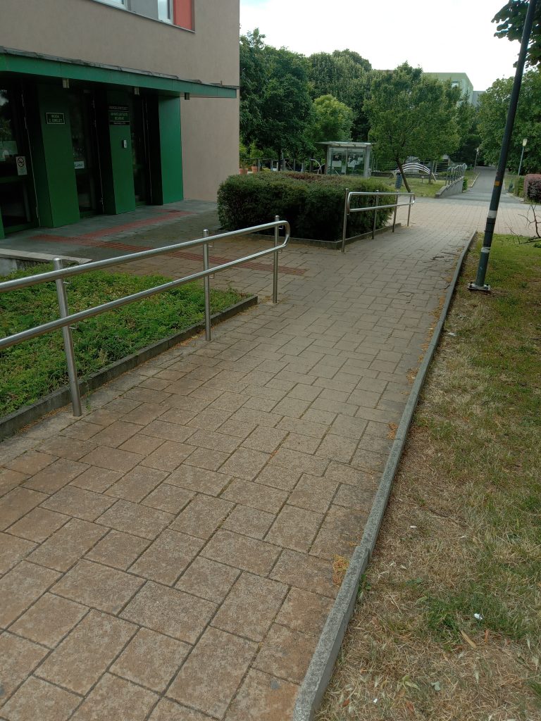 A rámpát látjuk, sima betonkocka burkolat, egyik oldalán korlát, nagyon enyhe emelkedővel.