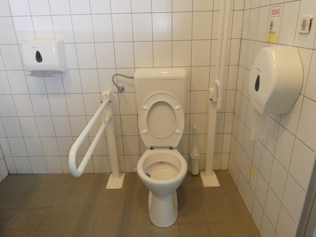 Az akadálymentes WC belülről. Belépve jobbra található a WC, mindkét oldalán felhajtható kapaszkodókkal.