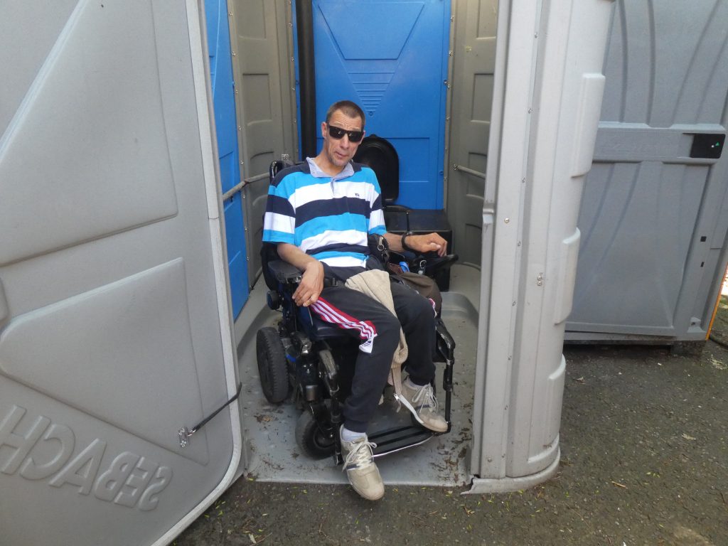 A mobil WC ajtajában Balázs áll elektromos kerekesszékkel. Látható, hogy simán befér.