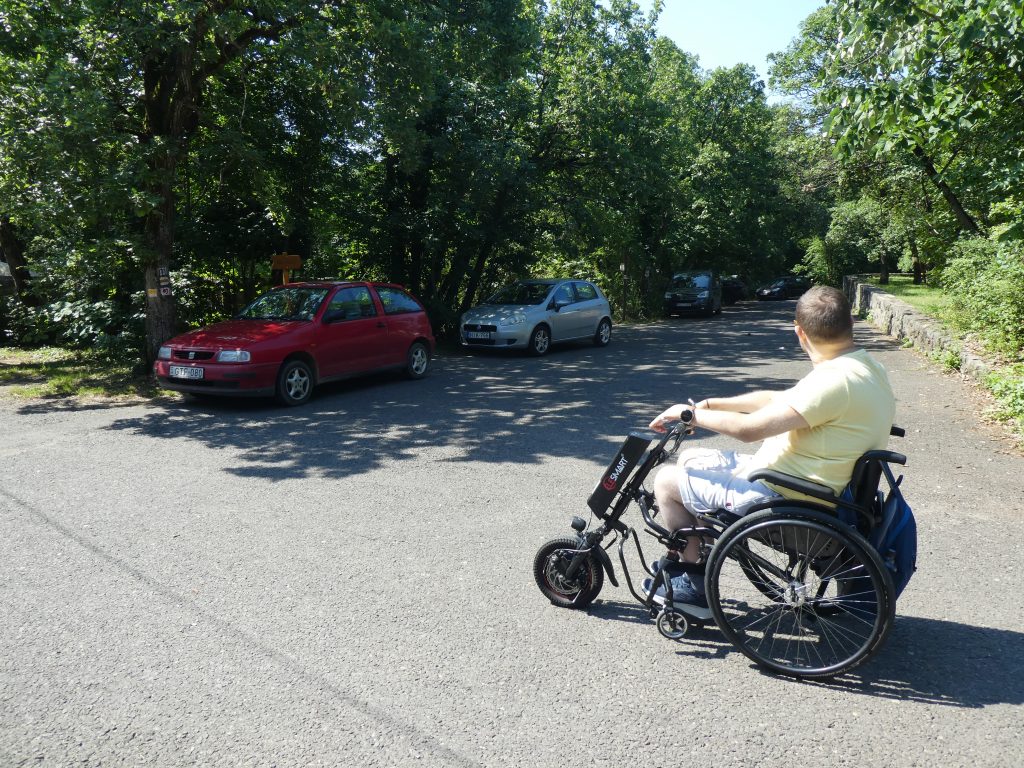 A parkolóban, a WC-k felé vezető útszakaszt látjuk, előttünk Gergő áll kerekesszékben. A parkoló árnyékos, sima betonborítású.
