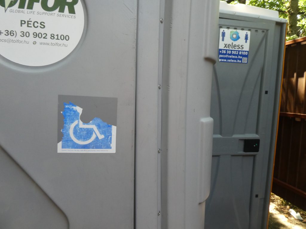 Két toi toi WC-t látunk egymás mellett illetve mögött. Az első, a baloldali akadálymentes. A WC-k fából készült paravánnal vannak körbevéve.