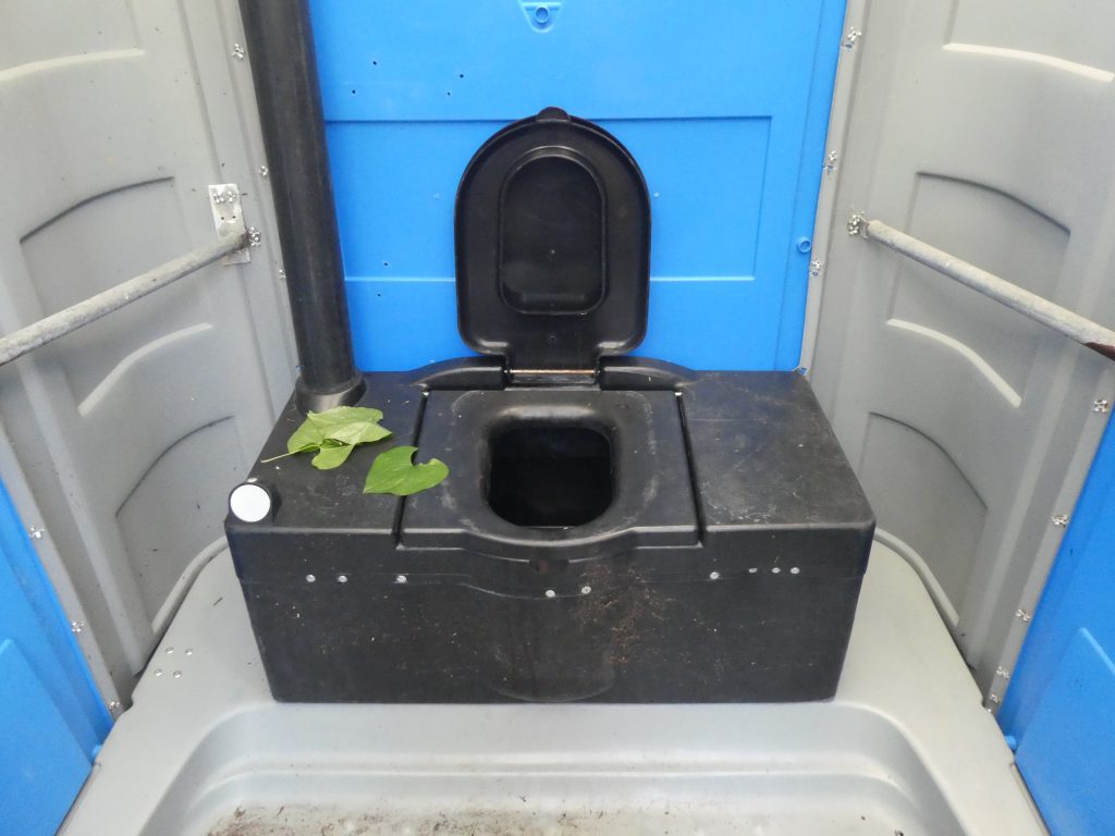 A mobil WC ülőkéjét fotóztuk közelről. Széles WC ülőke, középen maga a nyílás. Fedele is van. Kapaszkodó látható mind a két oldalon a falon, ám elég messze vannak a kényelmes használathoz. WC papírt nem látunk.