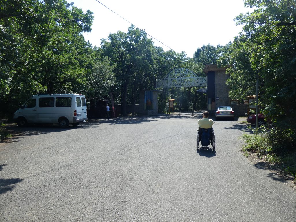 A bejárat távolról. Látszik, hogy nemcsak az oldalsó parkoló áll rendelkezésre, de 4-5 autó közvetlen a bejárat mellett is tud parkolni. A talaj sima betonút. A bejárat egy hatalmas kapu, küszöb, lépcső, ajtó nincsen.