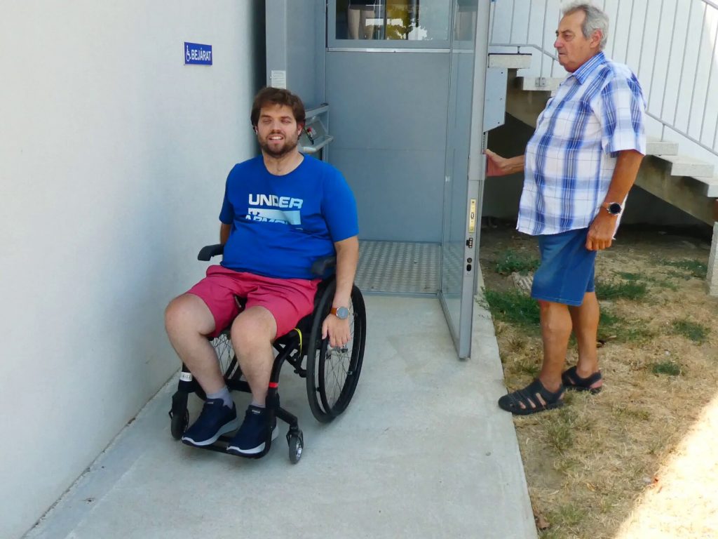 A külső liftből száll ki Gergő kerekesszékkel, mellette a sportcsarnok munkatársa nyitja neki a liftajtót. 