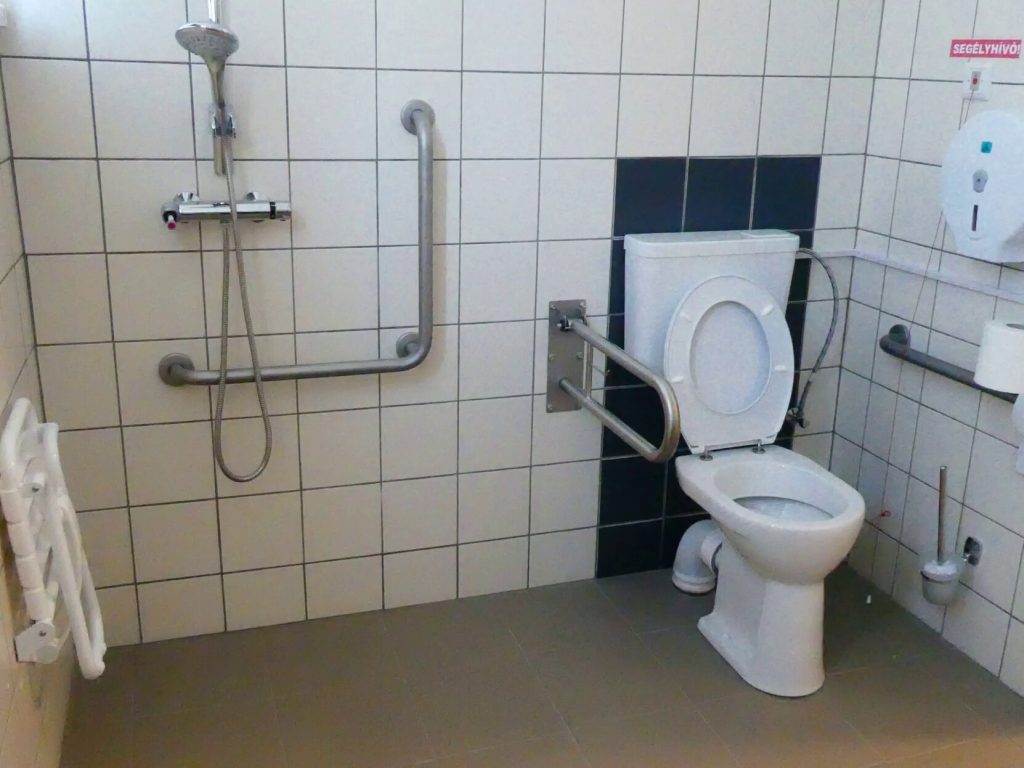 Az akadálymentes mosdó WC-je a képen, mely a helyiség jobb hátsó sarkában található. A falon rögzített, a másik oldalon térbe nyúló kapaszkodót látunk. A WC másik oldalán a falon kapaszkodóval ellátott, fülke és függöny nélkül, perem mentes tusoló található, a falra rögzített ülőkével. 