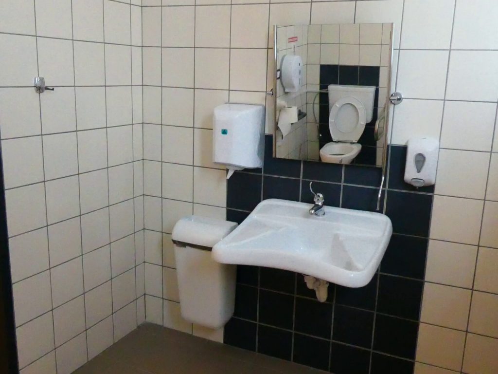 Az akadálymentes WC mosdója a képen, kerekített széléhez könnyen odaférni kerekesszékkel. Fölötte tükör. Balra a papíradagoló, jobbra a szappanadagoló található a falra rögzítve. 