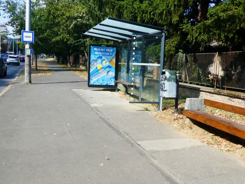 A Pétáv előtti buszmegálló. Széles járdán, sima út. Nem látni lekerekített széleket. A buszmegálló elejében esőbeálló paddal. Mellette kuka és egy újabb pad. 