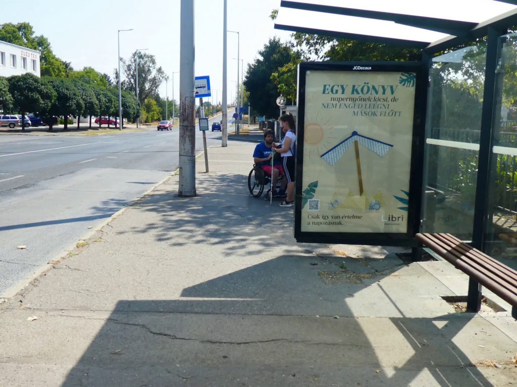 A Kertváros felé vezető buszmegálló a Pétávnál. Régi betonjárda, lekerekített széleket nem látunk. Középen esőbeálló paddal. 