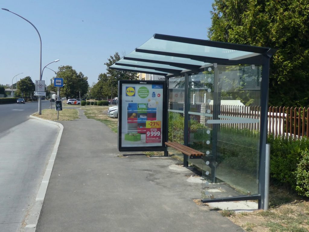 A Sportcsarnok buszmegálló Uránváros felé. Sima, új betonjárda, nem látunk lekerekített széleket. Középen esőbeálló található paddal. 