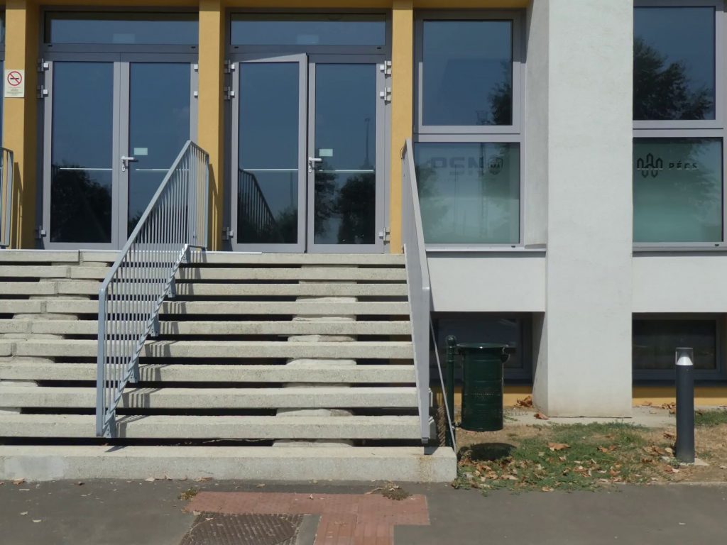 A lépcsős bejárat a képen, 10 lépcső vezet a széles lépcsősoron a bejárathoz. A kétszárnyú ajtók kifelé nyílnak. 