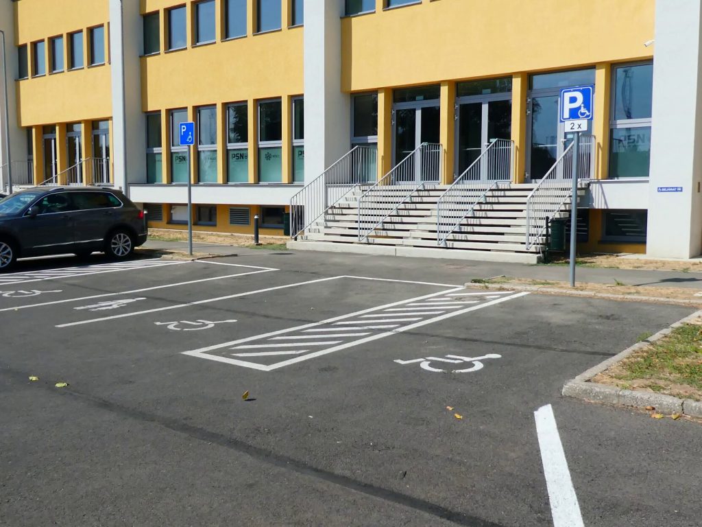 Szemből az épület elején lépcsősorokon közelíthető meg a bejárat. Ezek előtt három akadálymentes parkolót látunk. 