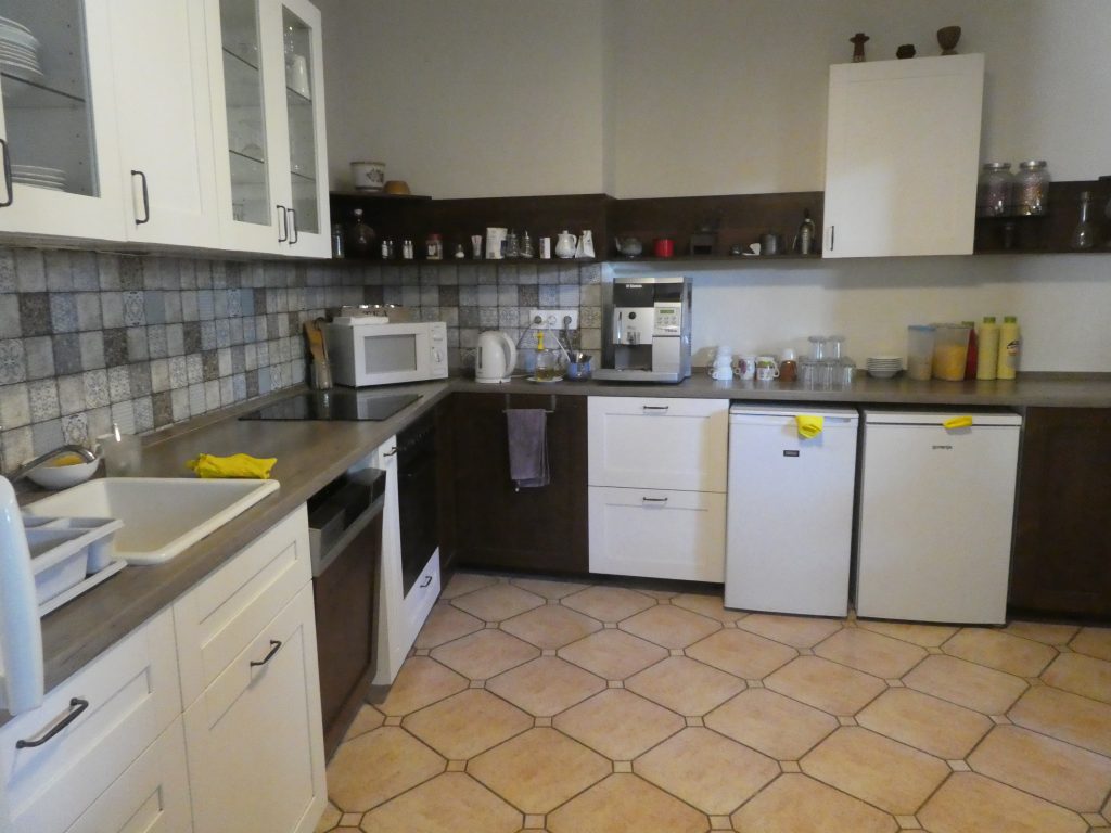 A közösségi térből nyíló konyha. L alakban található a konyhapult és az eszközök. Sima kő burkolatú, küszöbök nincsenek.