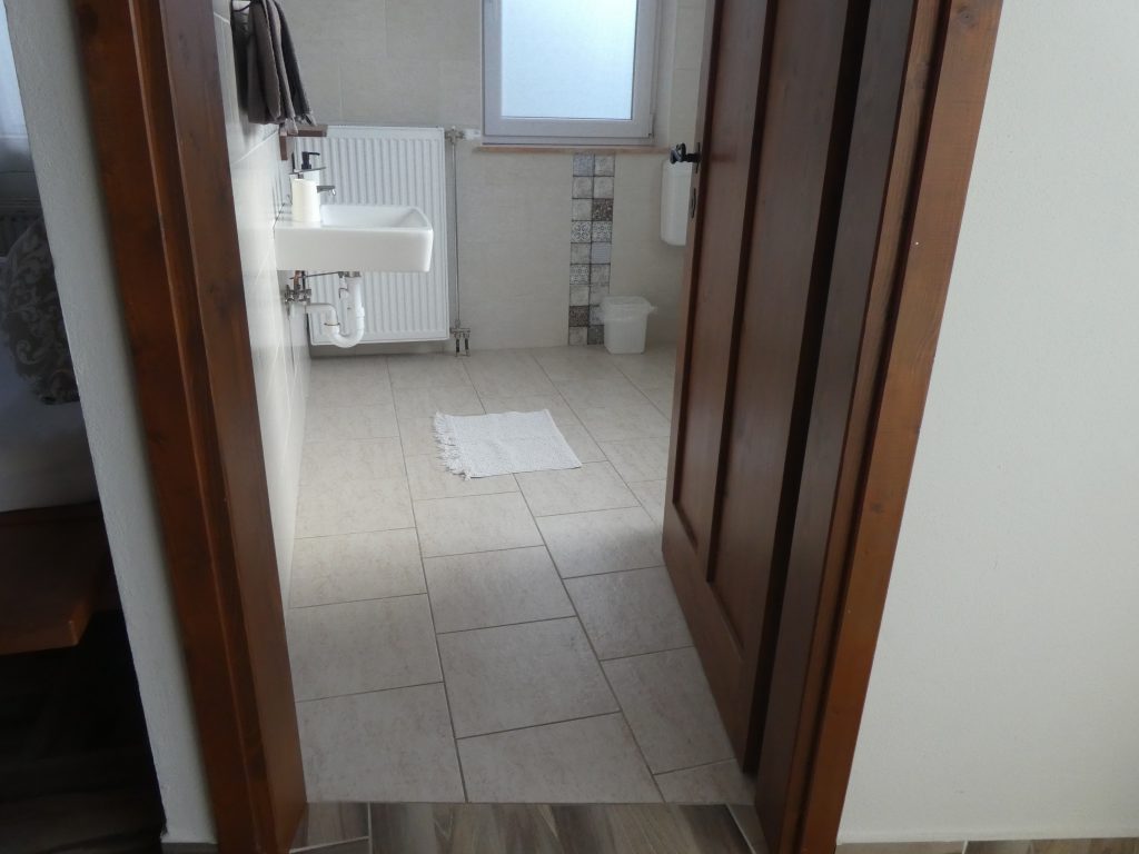 A fürdő ajtajában állunk. Küszöb nincsenek, folytatódik a sima kőburkolat. Balról találjuk a kézmosót, jobbról a tusolót, azzal szemben jobbra hátul a WC. Az ajtó befelé nyílik.