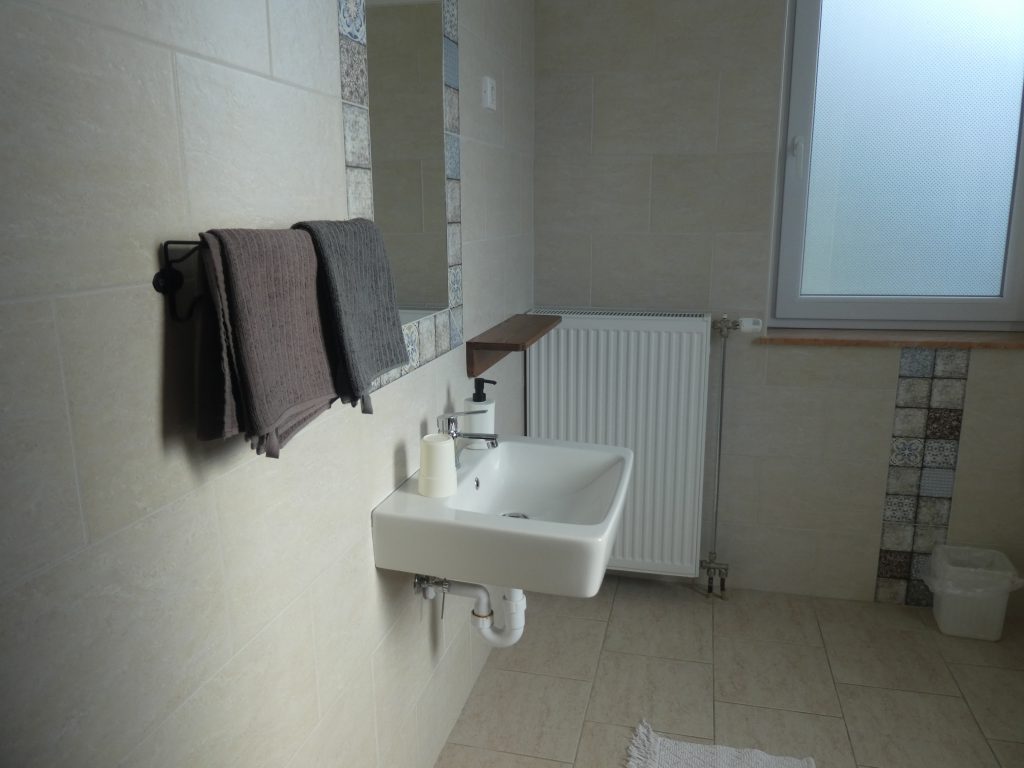 A fürdőszobába belépve balról található a kézmosó. A fürdő nagyméretű, sima kőburkolattal. Küszöbök nincsenek.